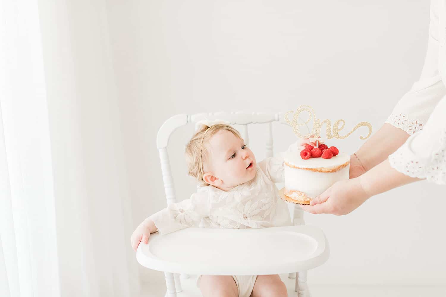 One-year-old baby girl in a white high chair looking at her first birthday cake during a baby photography session in Destin FL