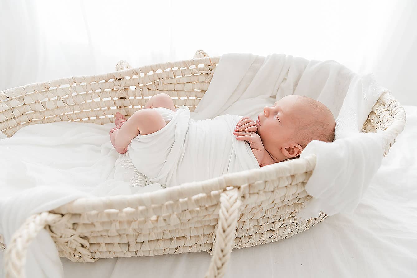 A newborn baby peacefully resting in a wicker basket from Coastal Crib Rentals.
