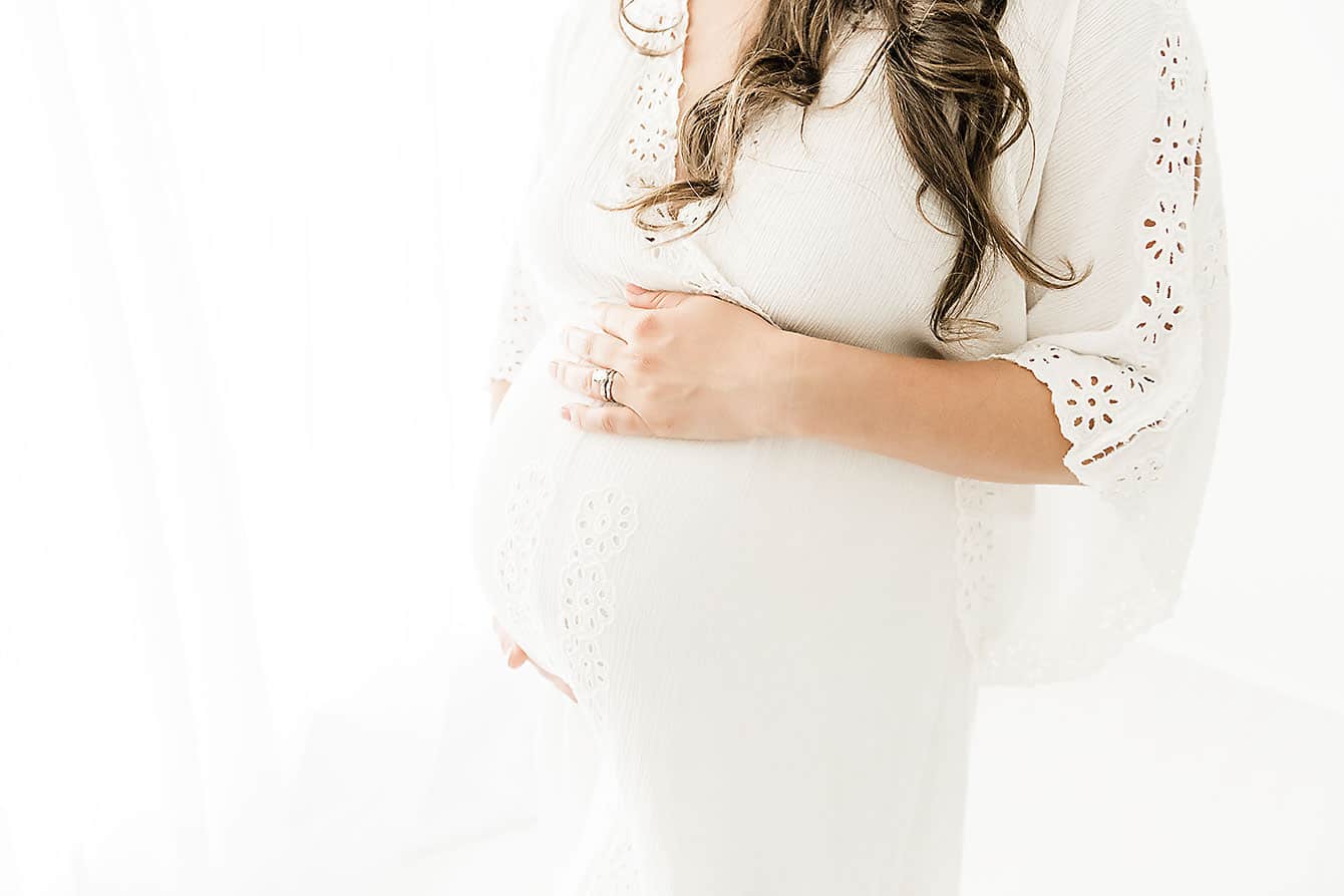 A pregnant woman in a white dress is posing for a photo at an OBGYN clinic in Pensacola, FL.