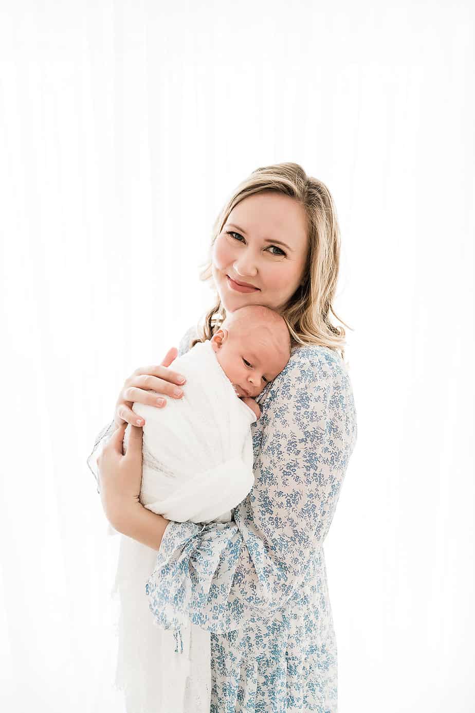Woman in a blue floral dress cradles her newborn, against a bright background.