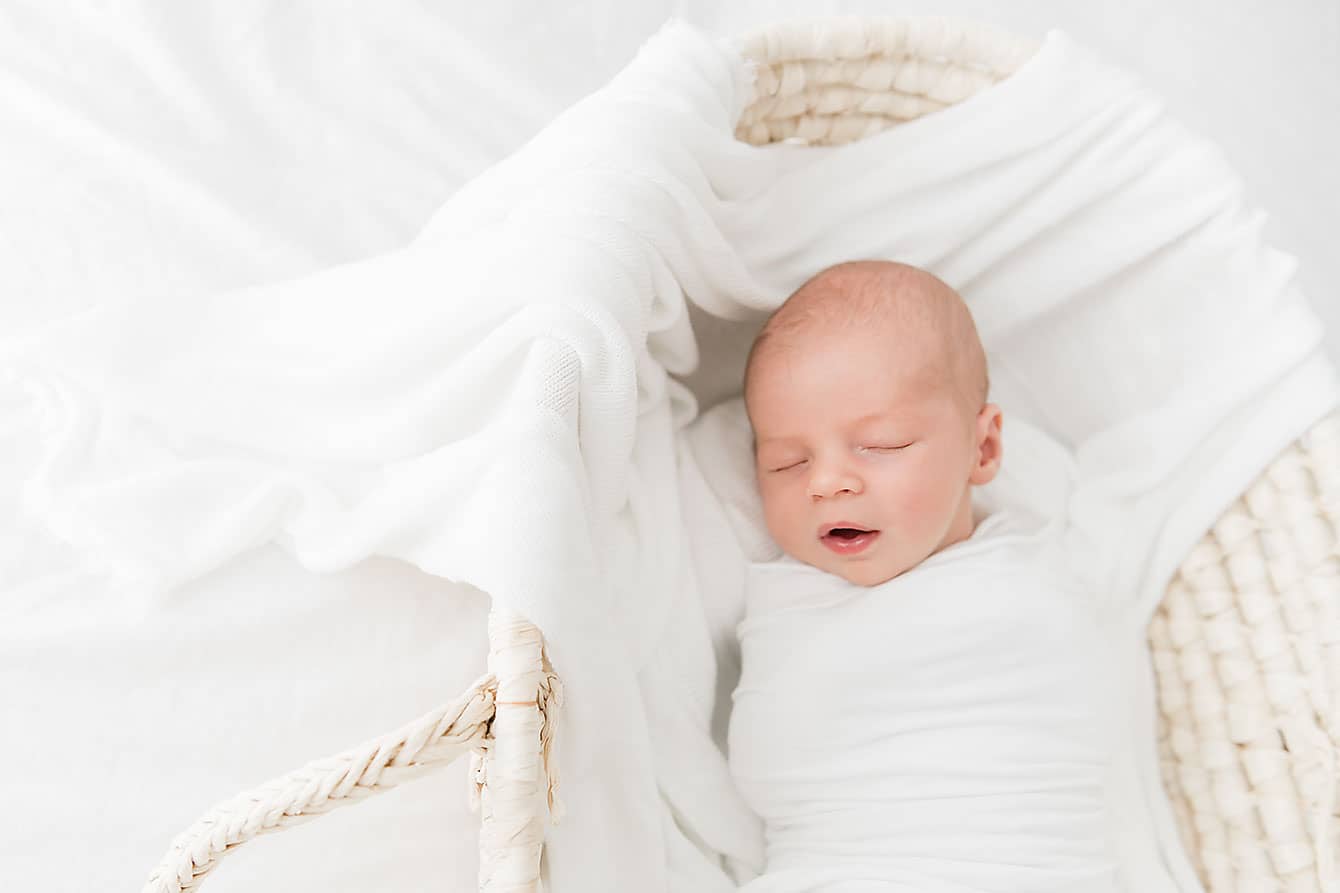 A newborn baby is swaddled in white fabric, sleeping peacefully in a woven basket on a white background.