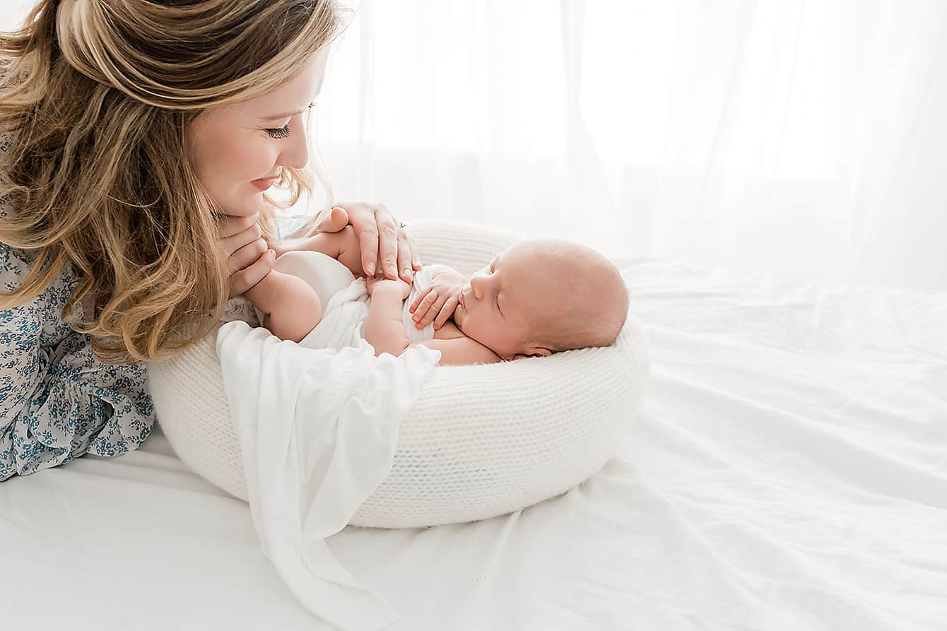 A woman gently leans over a sleeping baby wrapped in a white blanket, lying on a bed bathed in soft, natural light, perfectly.