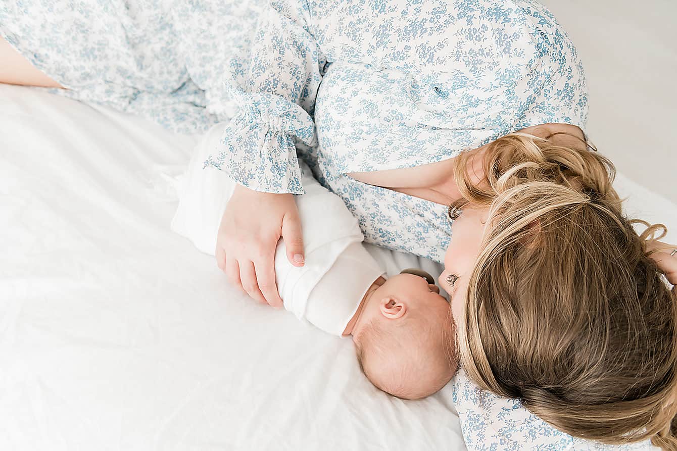 Woman lying on a bed breastfeeding her newborn, both in light attire—captured by a Niceville newborn photographer.