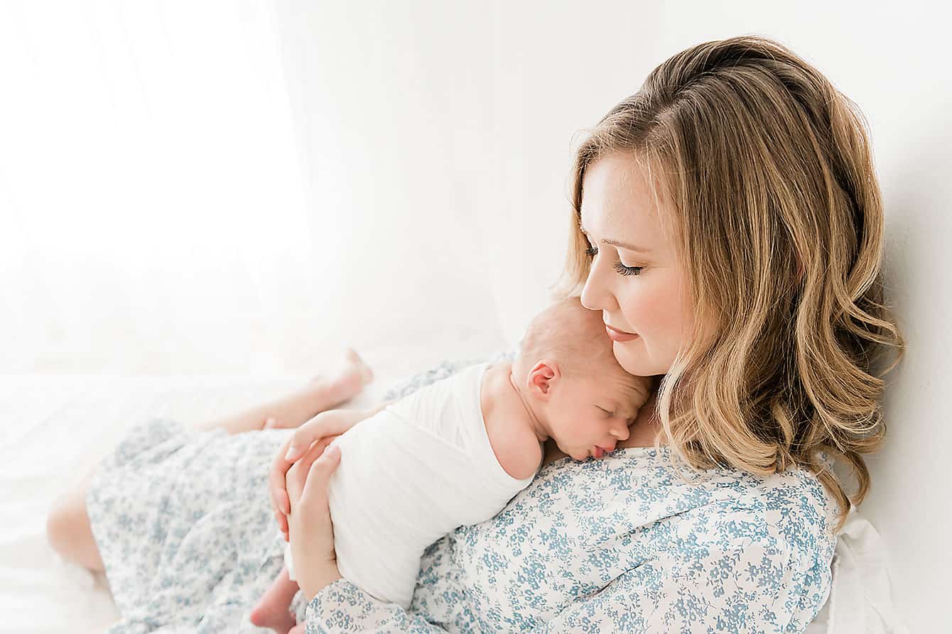 A woman with shoulder-length hair cradles a sleeping baby wrapped in a white blanket, captured beautifully by a Niceville newborn photographer. They are seated in front of a bright, softly lit background.