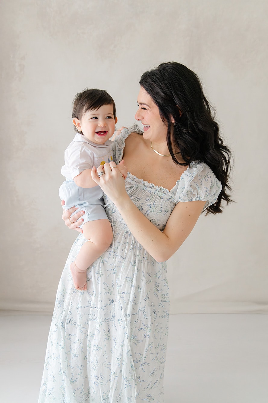 Woman in a light dress holds a smiling baby, styled like those from boutiques in Destin Florida, against a softly-lit background.