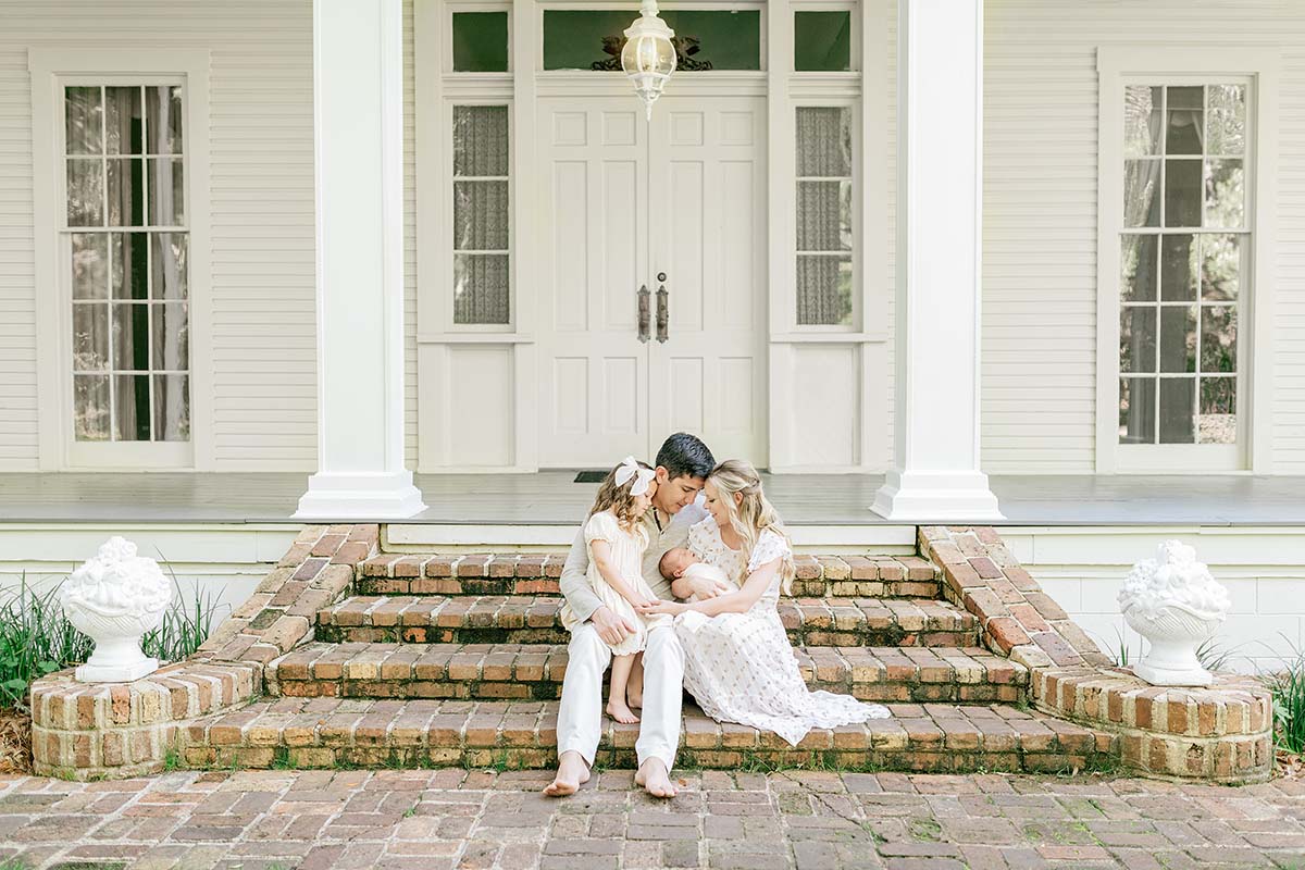 family of four with newborn and older sister sitting on brick steps of a white home photographed by a 30A newborn photographer