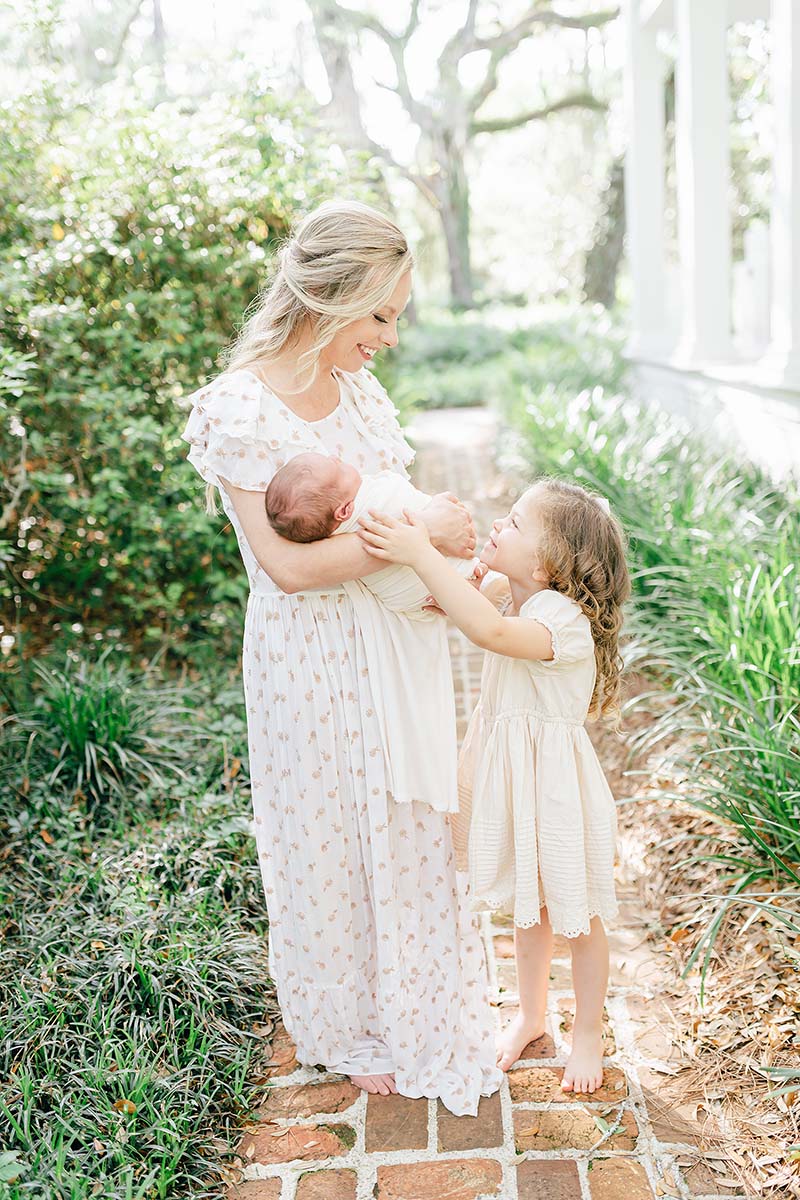 mother holding newborn while older sister reaches up to touch baby during an in-home newborn session