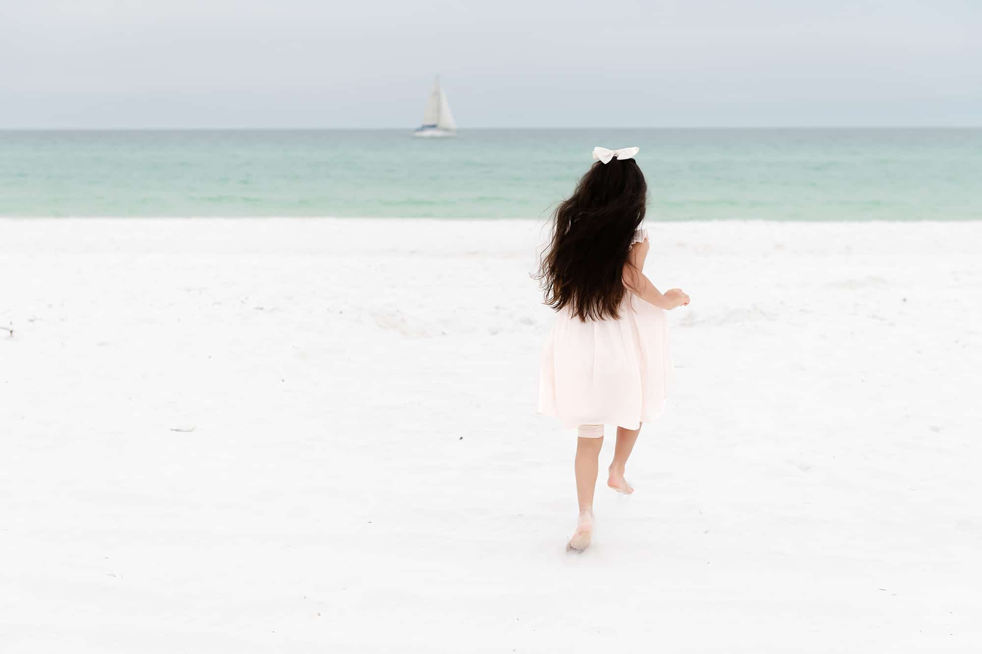 A young girl in a light dress runs barefoot on a white sandy beach near Pediatric Dentist Niceville, with a sailboat in the distance.