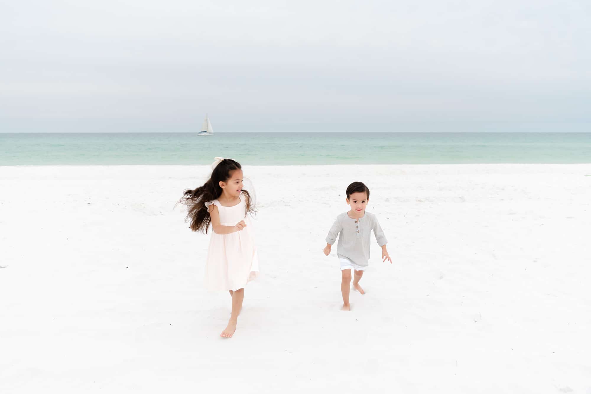 Two young children run barefoot on a white sandy beach near Niceville, smiling like patients of a pediatric dentist.