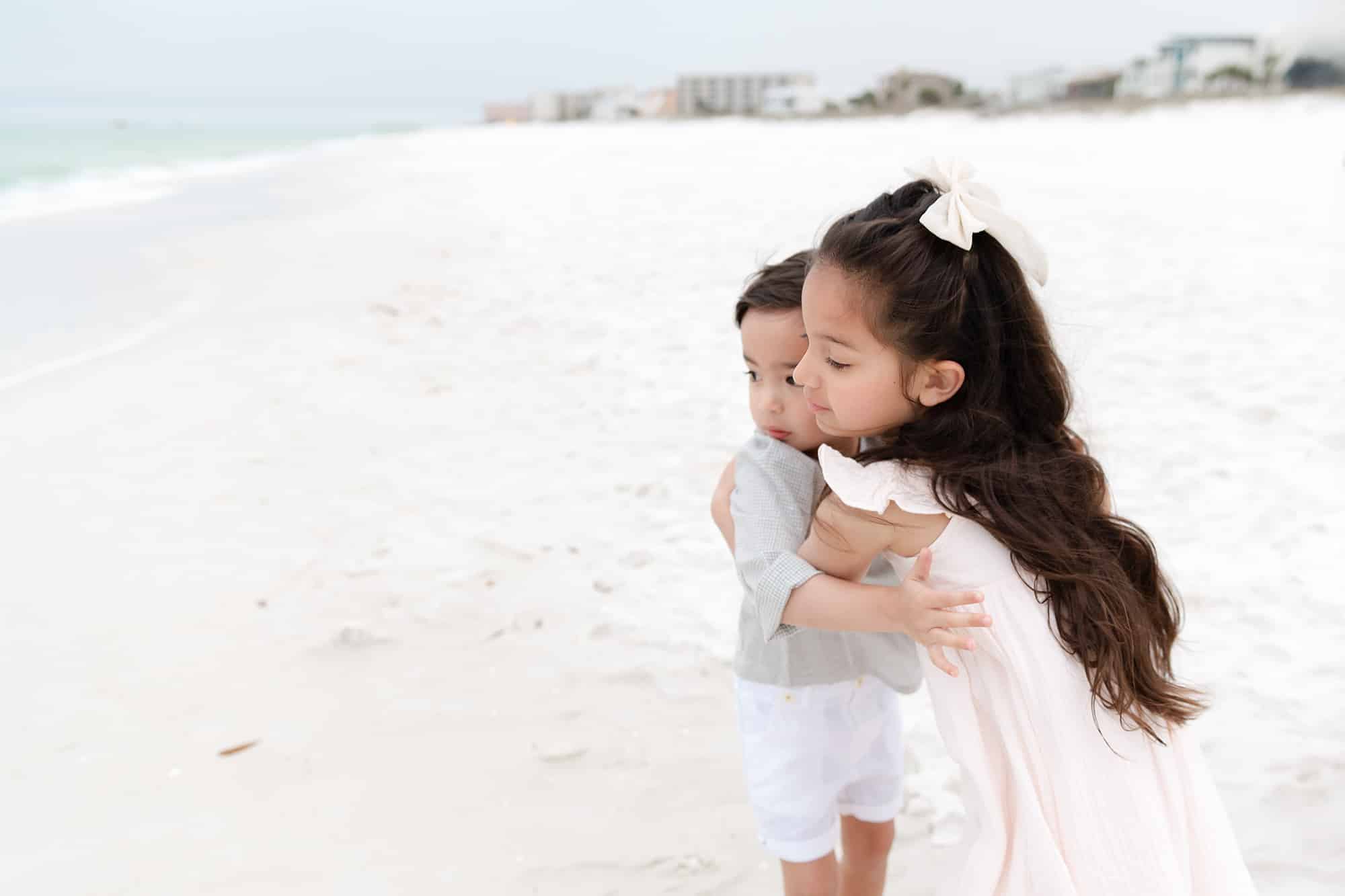 A young girl hugs a younger boy on a sandy beach near buildings, both smiling like they just left their Pediatric Dentist Niceville.