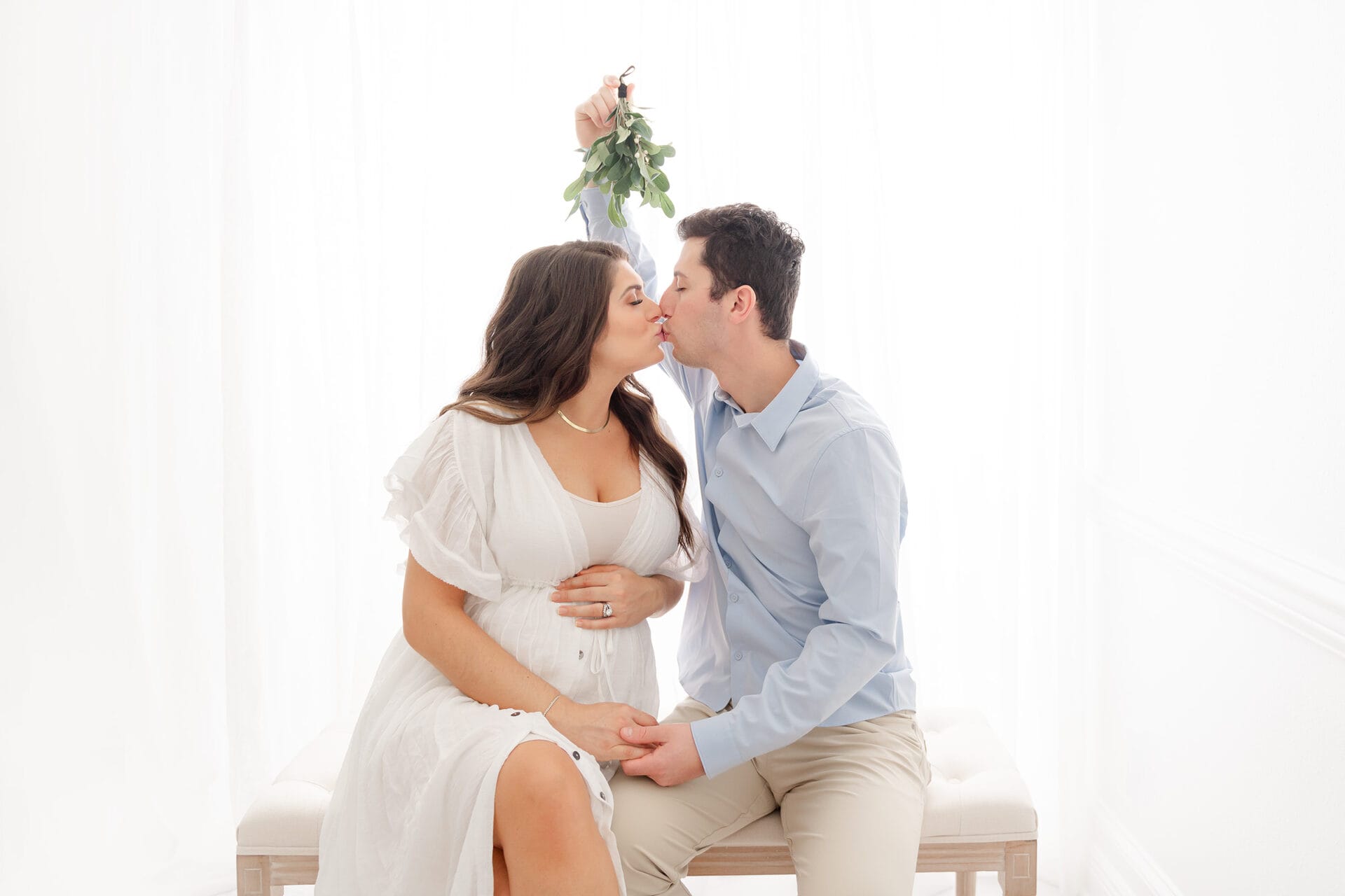 A Pensacola maternity photographer captures a tender moment of a pregnant couple kissing in front of a white background.