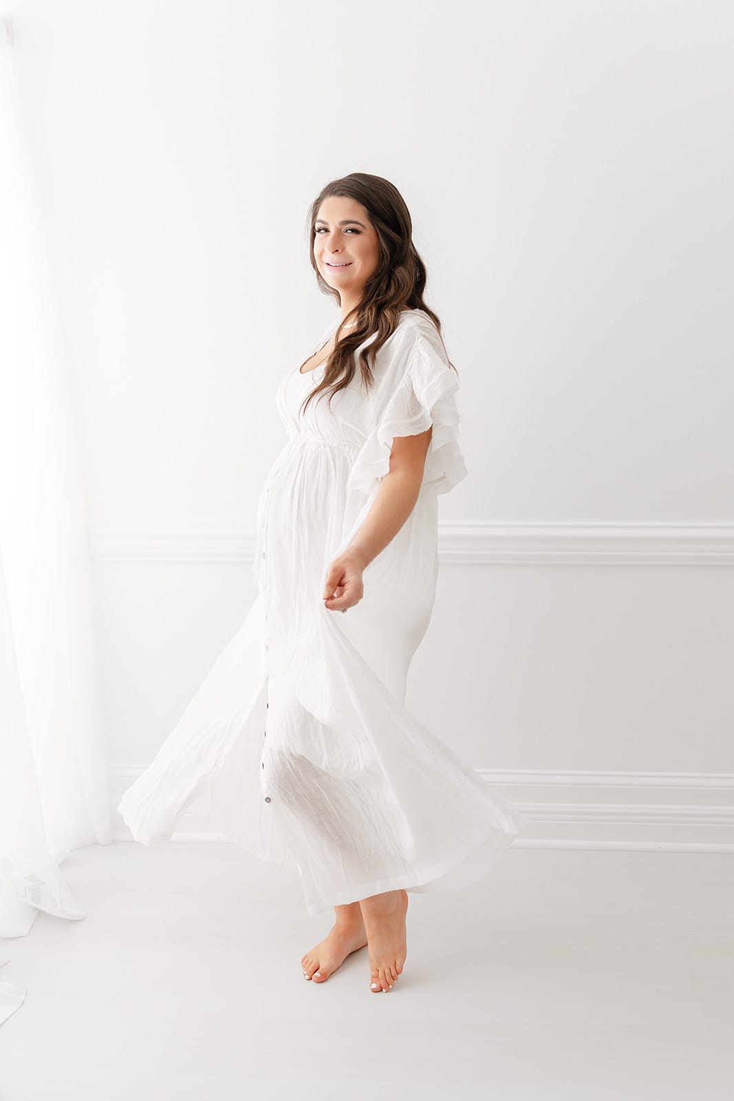 A Pensacola maternity photographer captures a pregnant woman in a white dress posing elegantly in front of a white wall.