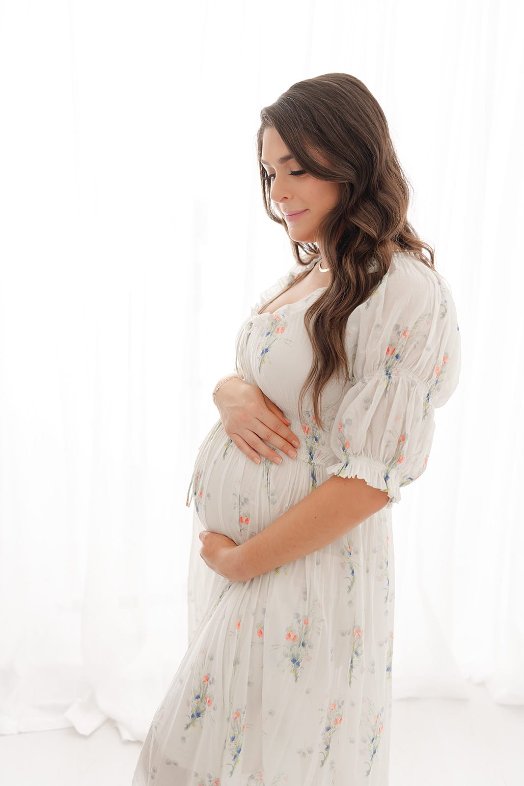 A pregnant woman in a floral dress poses in front of a white background, captured by a talented Pensacola maternity photographer.