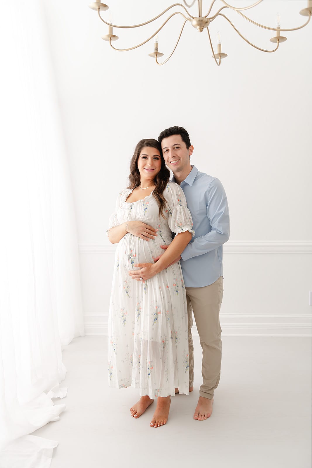 A Pensacola maternity photographer captures an expectant couple striking a pose in front of a chandelier in a pristine white room.