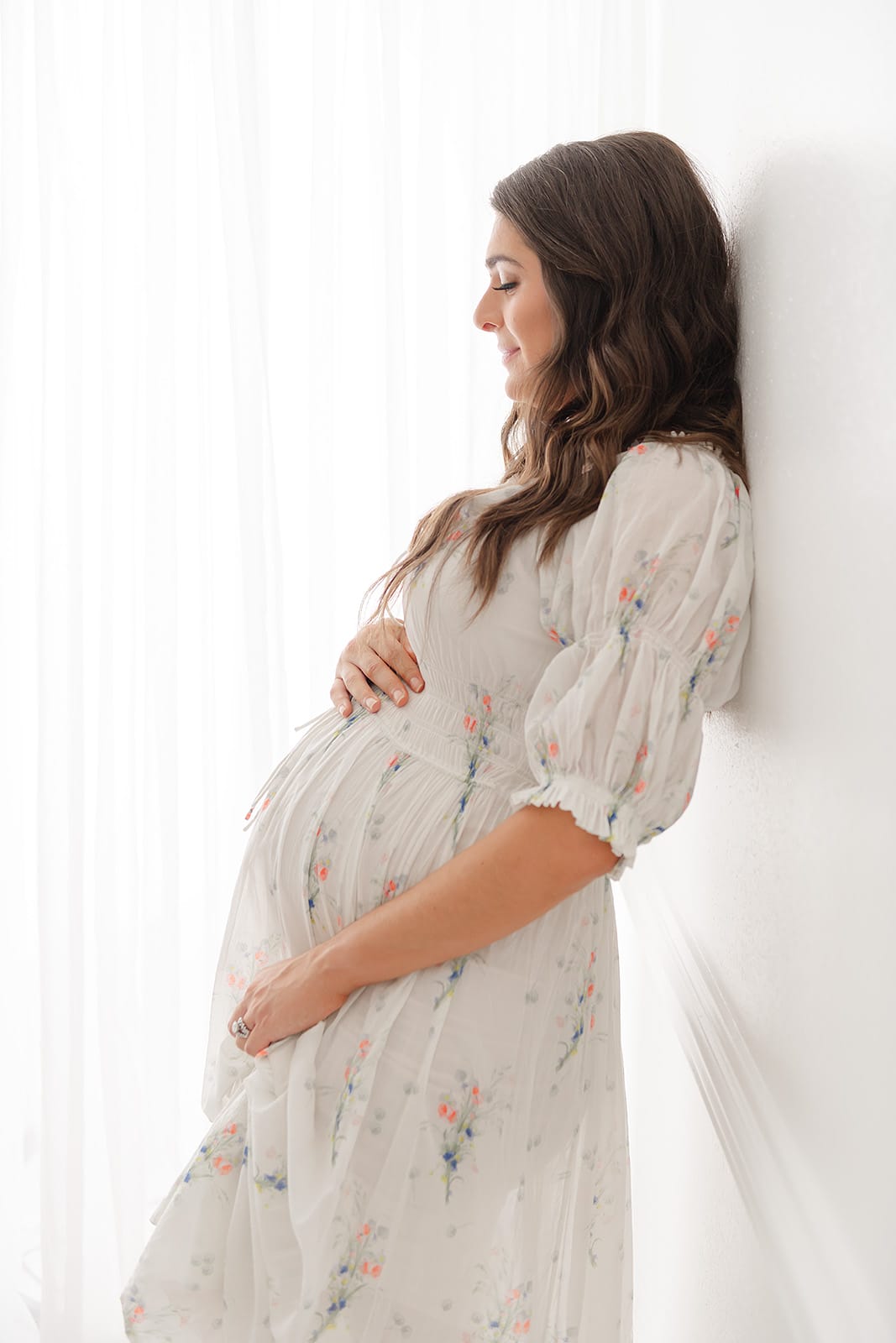 A Pensacola maternity photographer captures a pregnant woman in a floral dress leaning against a wall.