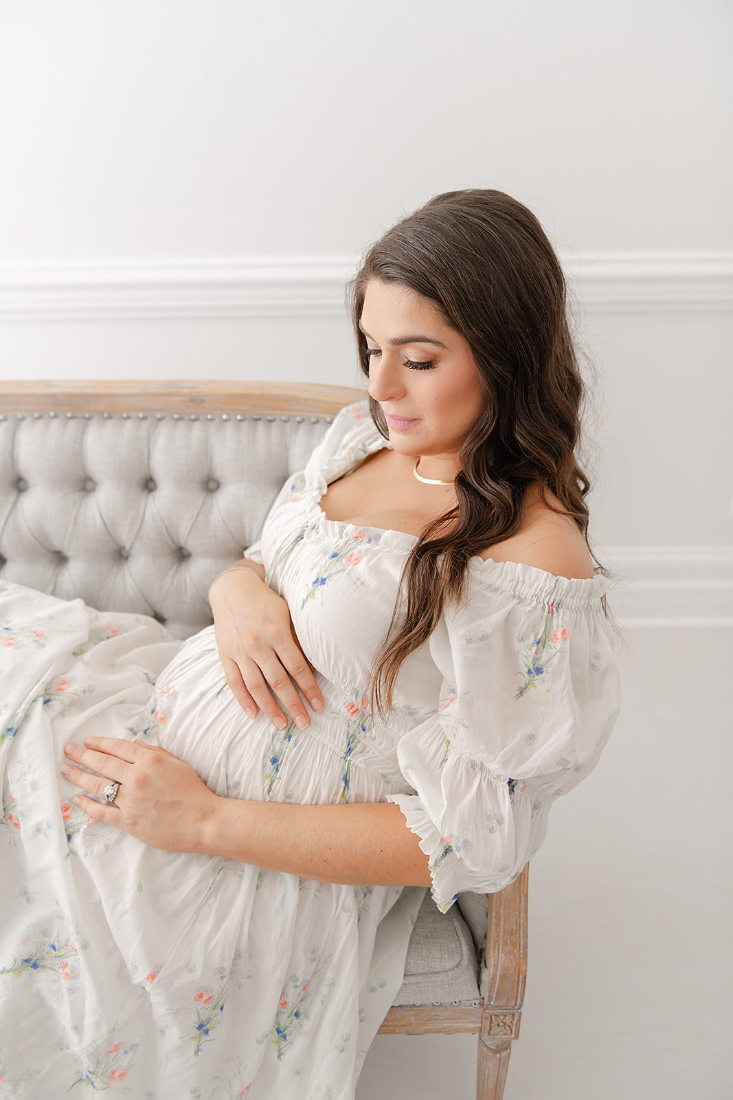 A pregnant woman in a floral dress, sitting on a gray couch.