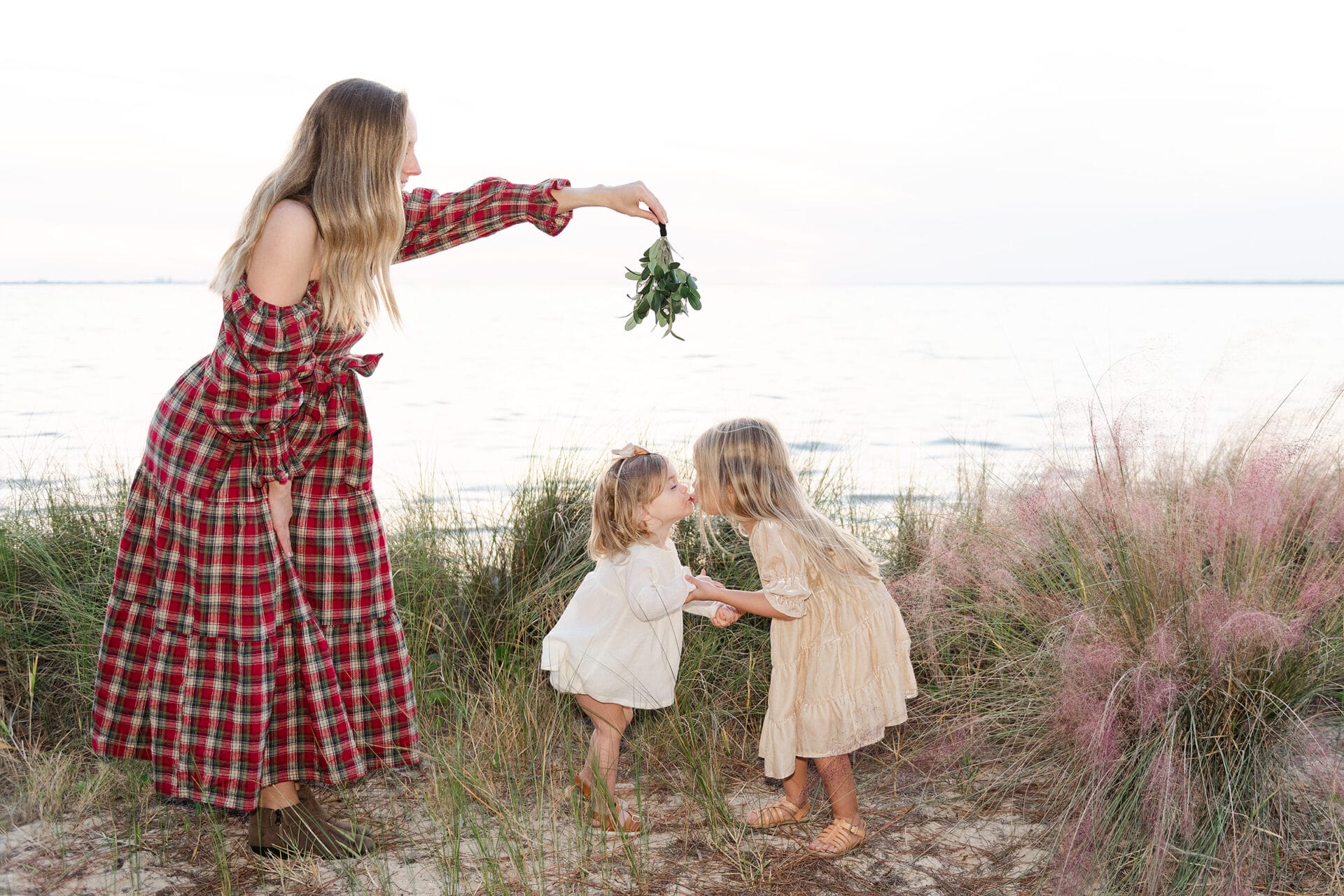 A woman in a plaid dress holds greenery above two young girls at the beach. The girls, in light dresses, stand in grassy sand by the water.