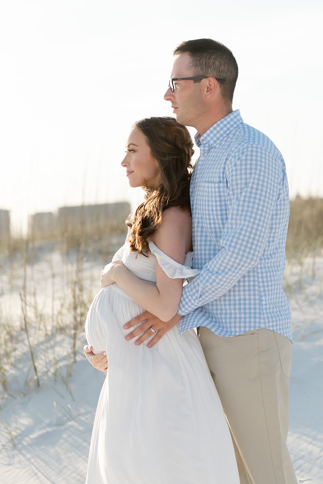 A pregnant woman in a white dress stands on a beach, embraced from behind by a man in a blue checkered shirt. They both gaze into the distance.