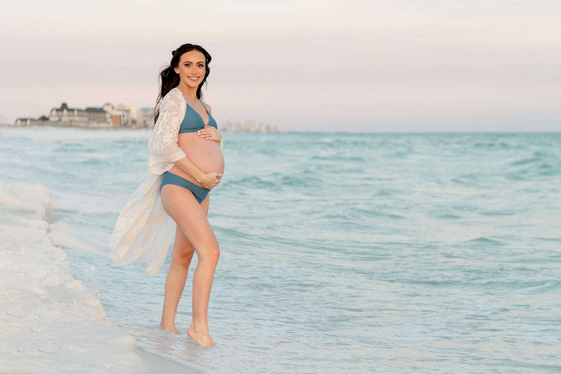 Pregnant woman stands on a beach, wearing a bikini and lace cover-up, with calm ocean waves and a distant shoreline in the background.