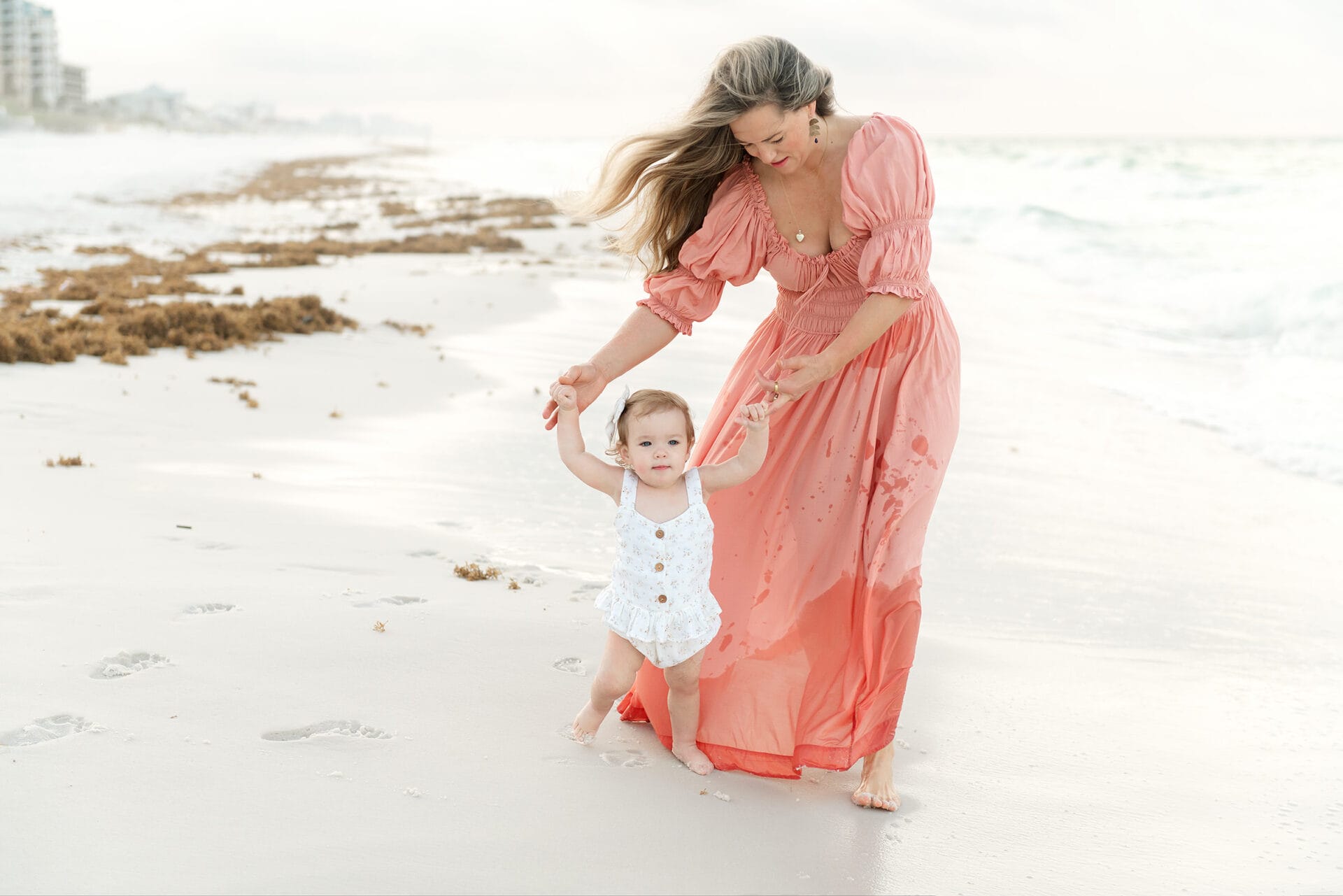 A woman in a pink dress guides a toddler in a white outfit along a sandy beach, enjoying a peaceful moment away from the bustle of private schools in Destin, FL.