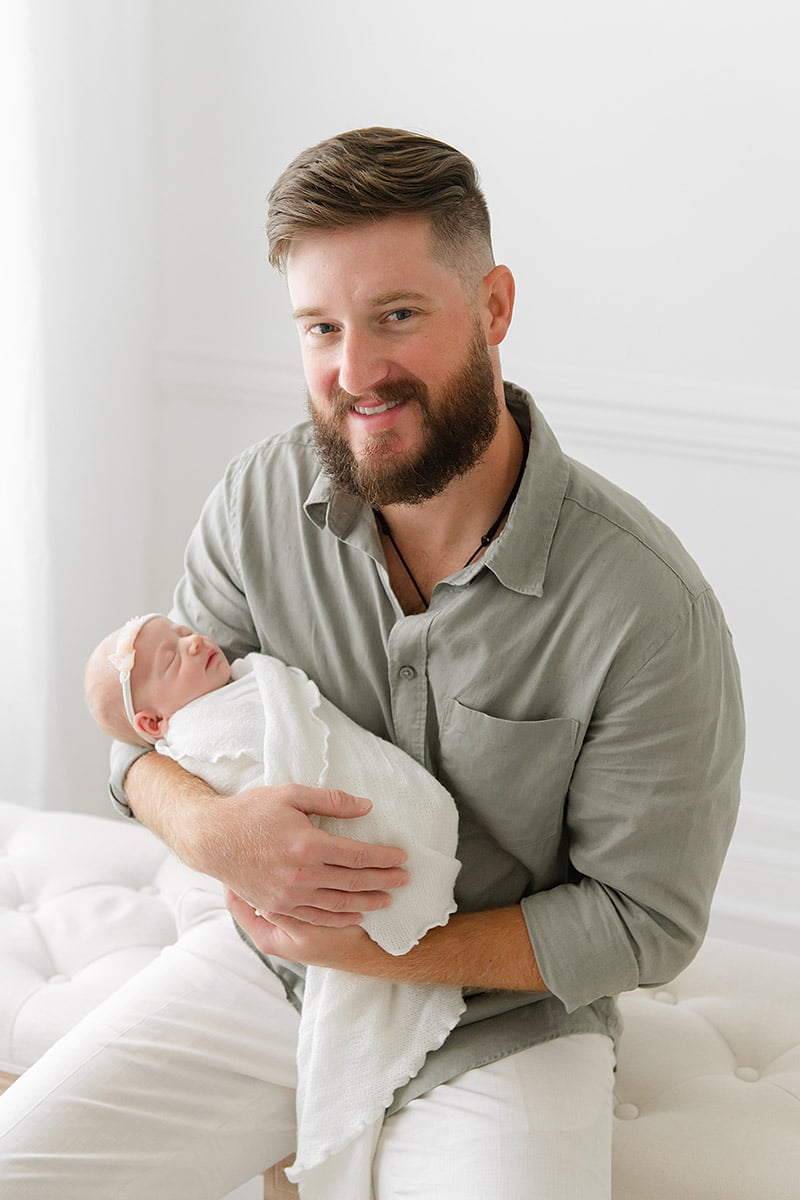 Proud new dad smiling and holding his swaddled newborn baby girl in a bright white studio
