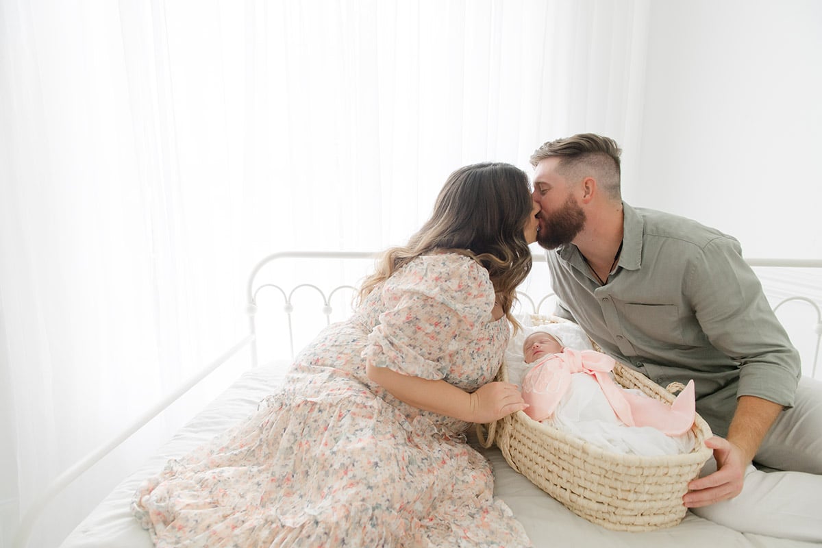 New parents sharing a kiss over their sleeping newborn in a wicker basket, photographed by a Fort Walton Beach newborn photographer at Andrea Krey Photography