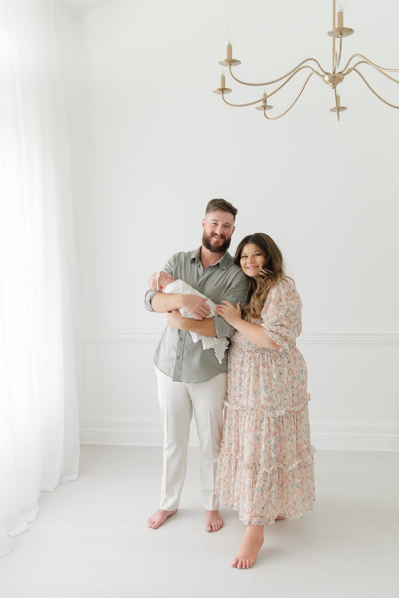 New family of three standing together in a bright white studio with a gold chandelier, photographed by Fort Walton Beach newborn photographer Andrea Krey