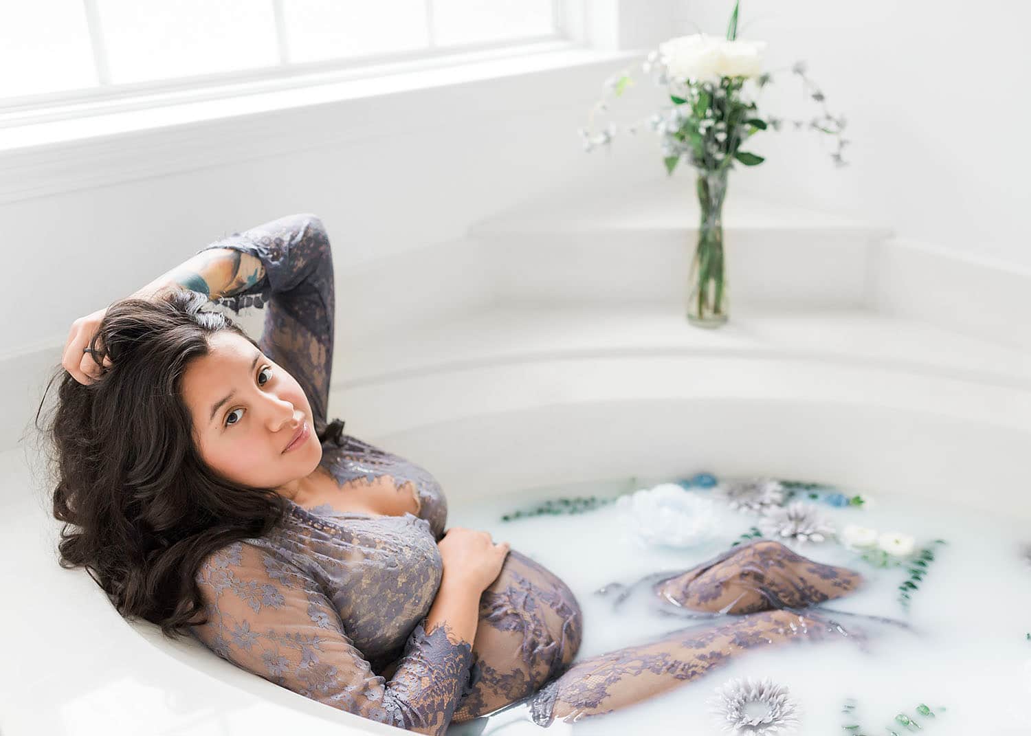 Pregnant woman in a purple lace gown relaxes in a bright milk bath with soft natural window light flooding the room