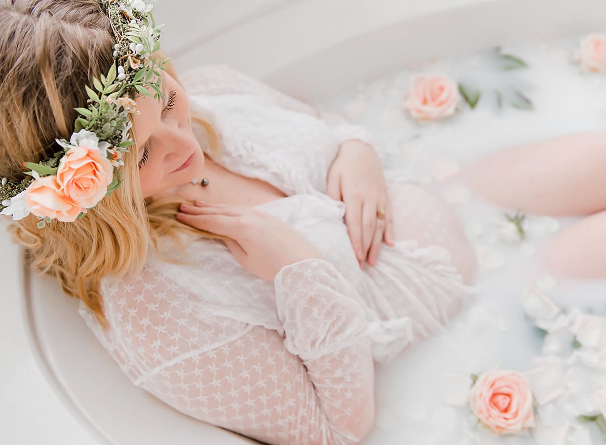 Expectant mother in a white lace robe reclines in a floral milk bath with peach roses during her milk bath maternity photos Destin FL session