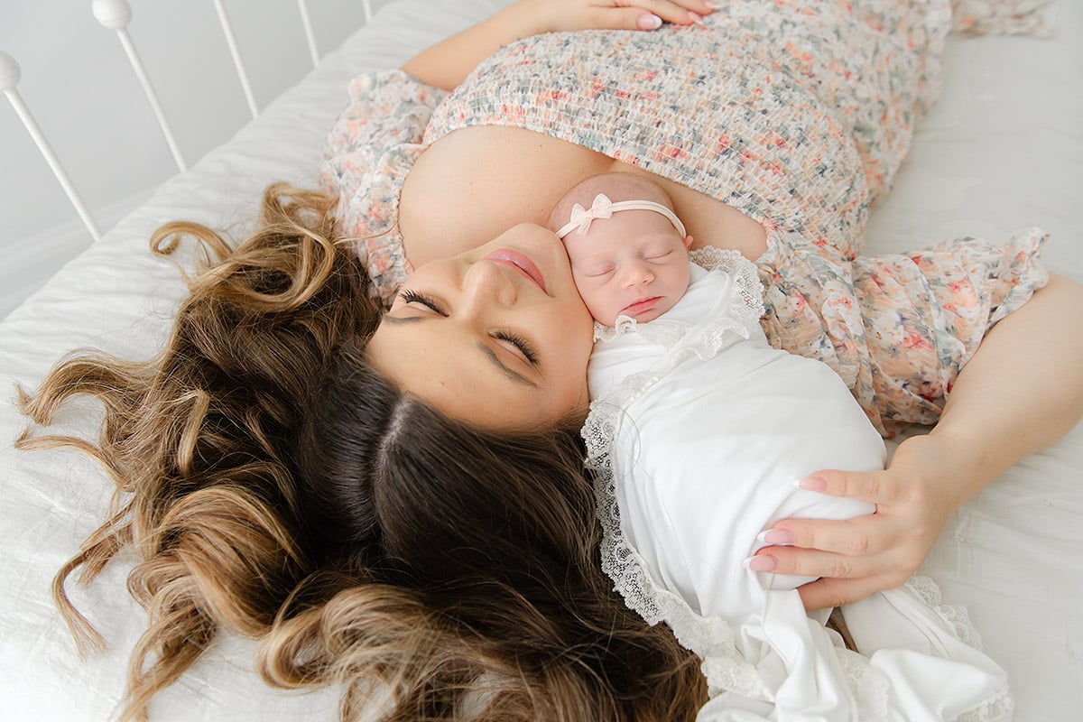 New mother lying cheek to cheek with her sleeping newborn baby, both dressed in soft florals and white, in a bright airy studio