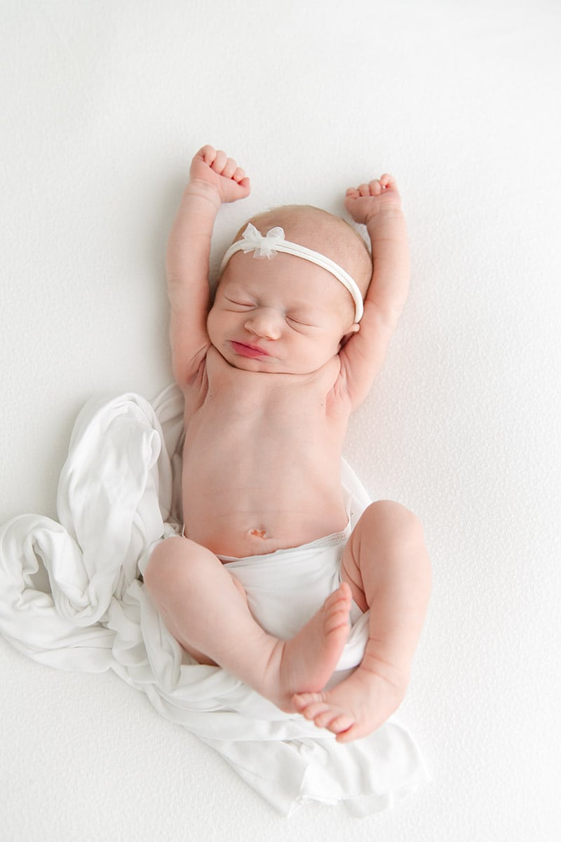 Newborn baby girl stretching her arms with a white bow headband, photographed on a clean white backdrop in a bright studio