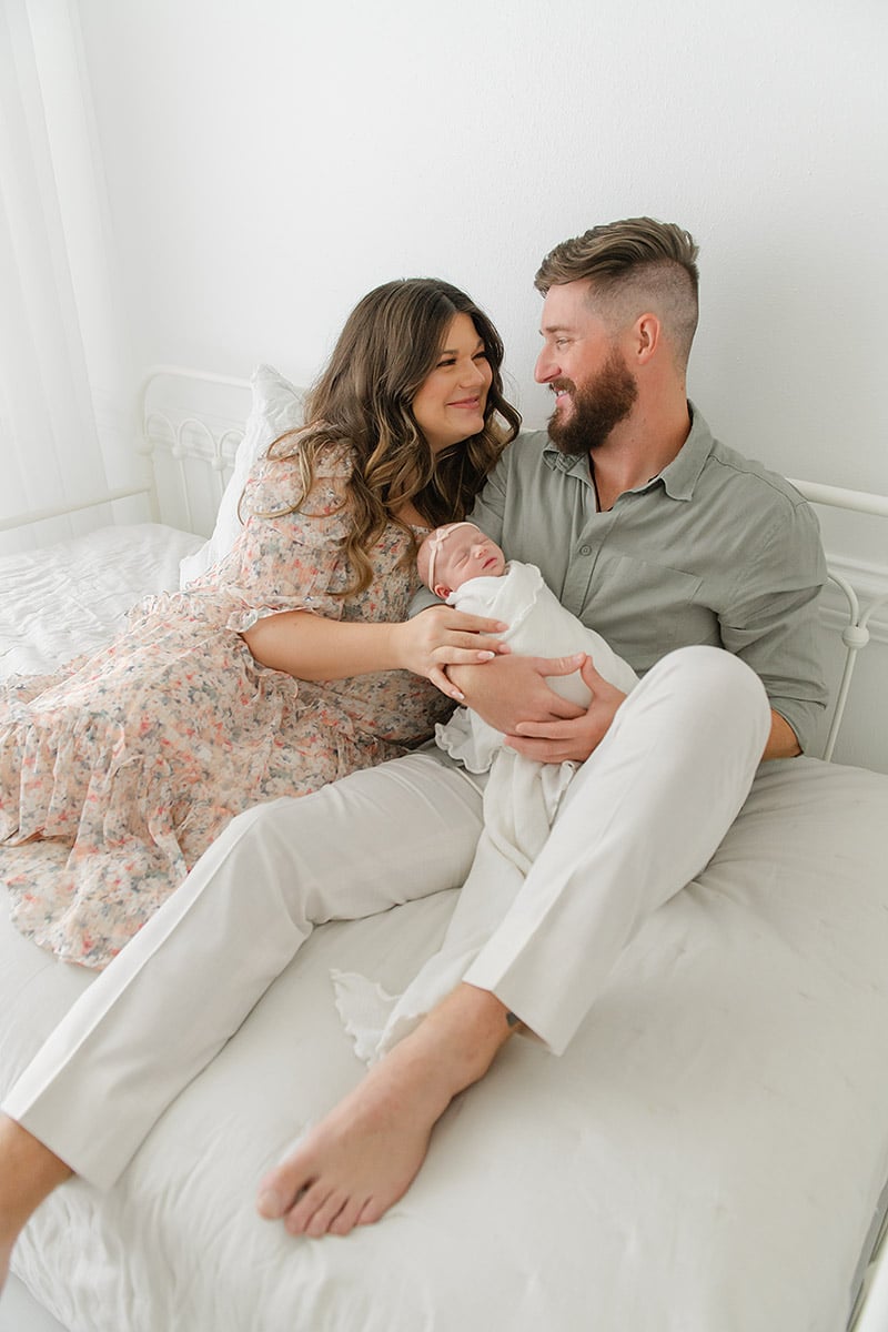 Happy new parents sitting together on a white bed smiling at each other while holding their newborn, photographed by a Fort Walton Beach newborn photographer