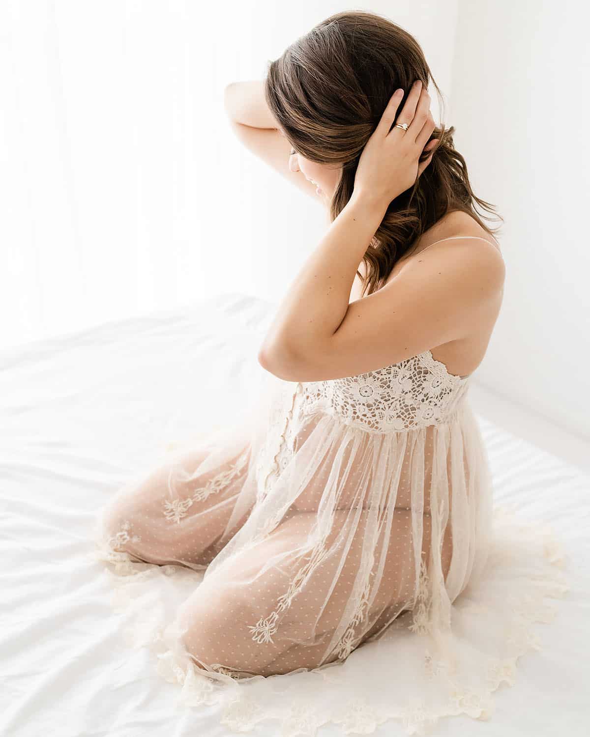 niceville-maternity-boudoir-portrait-sheer-lace-negligee-studio.jpg Alt text: Expecting mother in a sheer lace negligee seated on white bedding with her hand in her hair during a boudoir-inspired Niceville maternity studio portrait