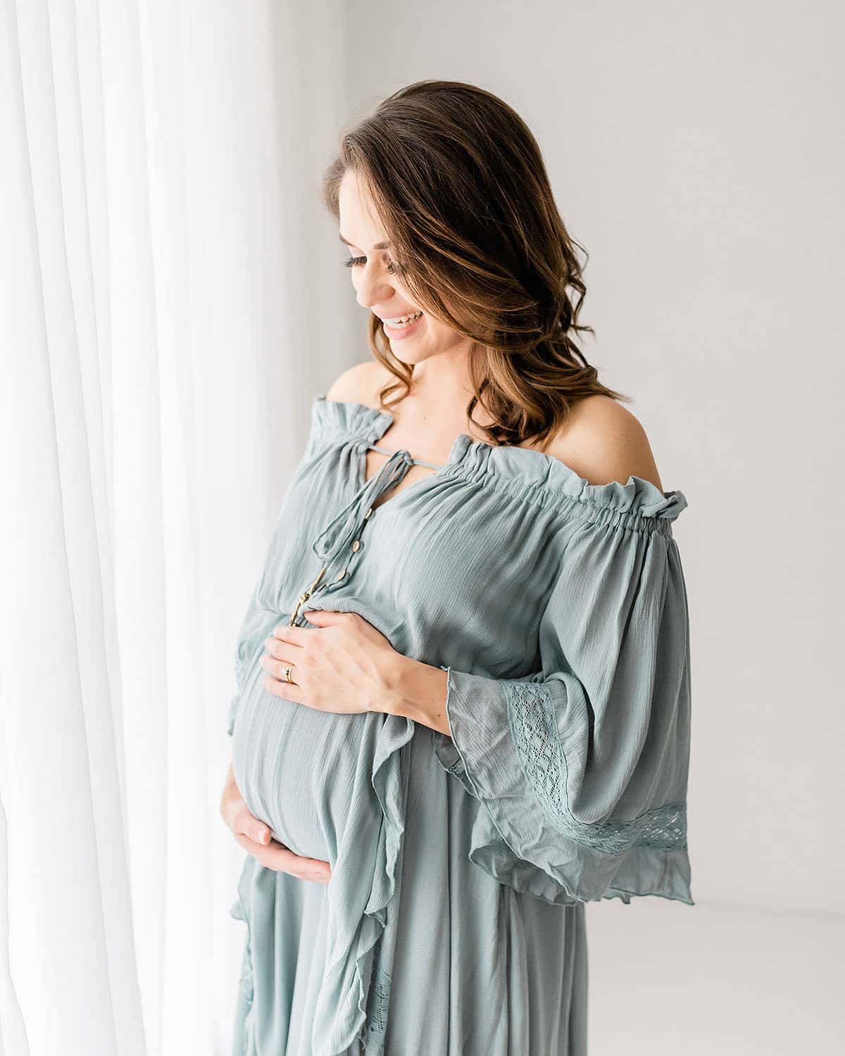 Expecting mother in a sage off-shoulder boho gown with lace sleeves smiling by a sheer curtain during a Niceville maternity photographer session