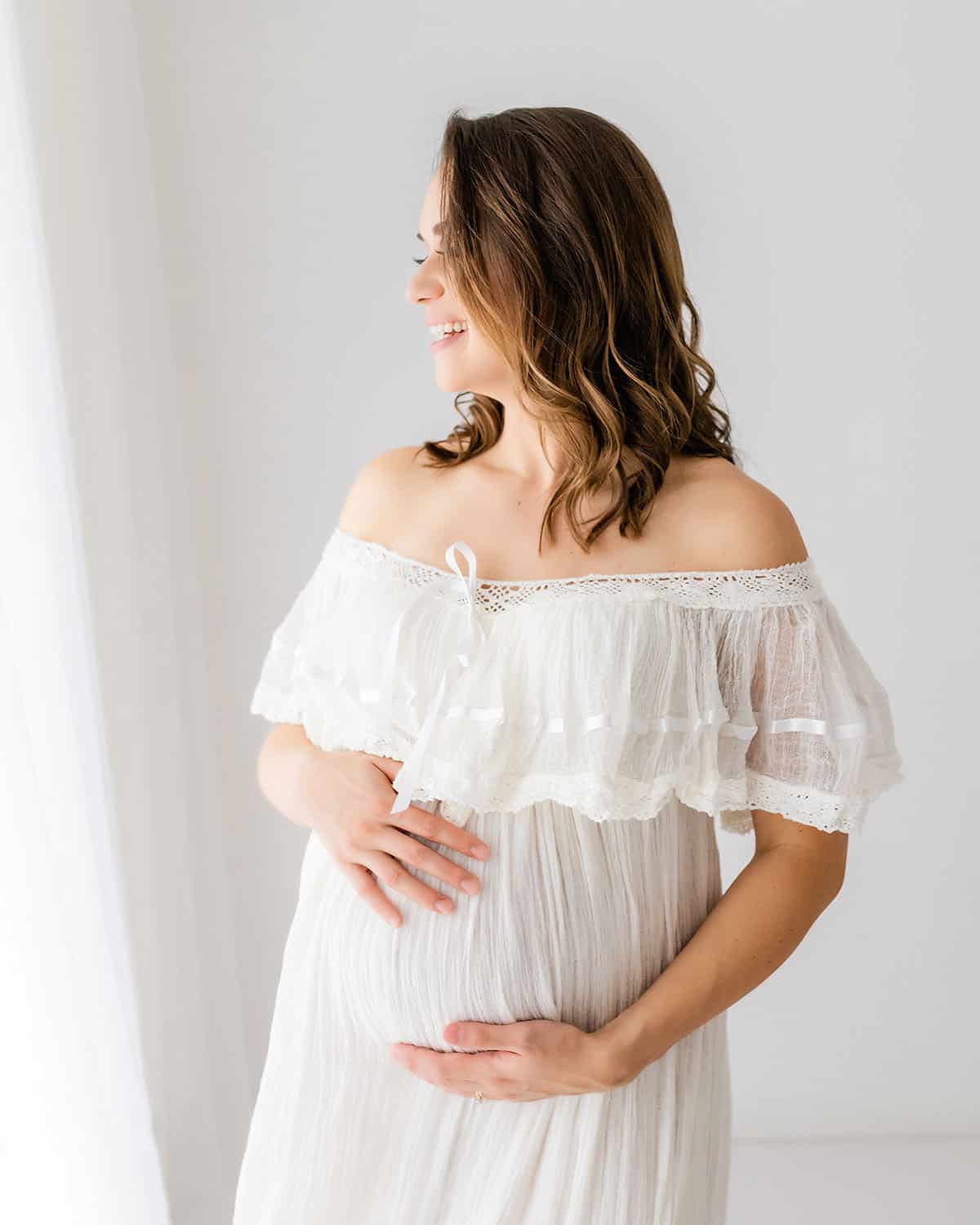 Glowing expecting mother in a white off-shoulder lace gown laughing joyfully during a Niceville maternity photographer studio session
