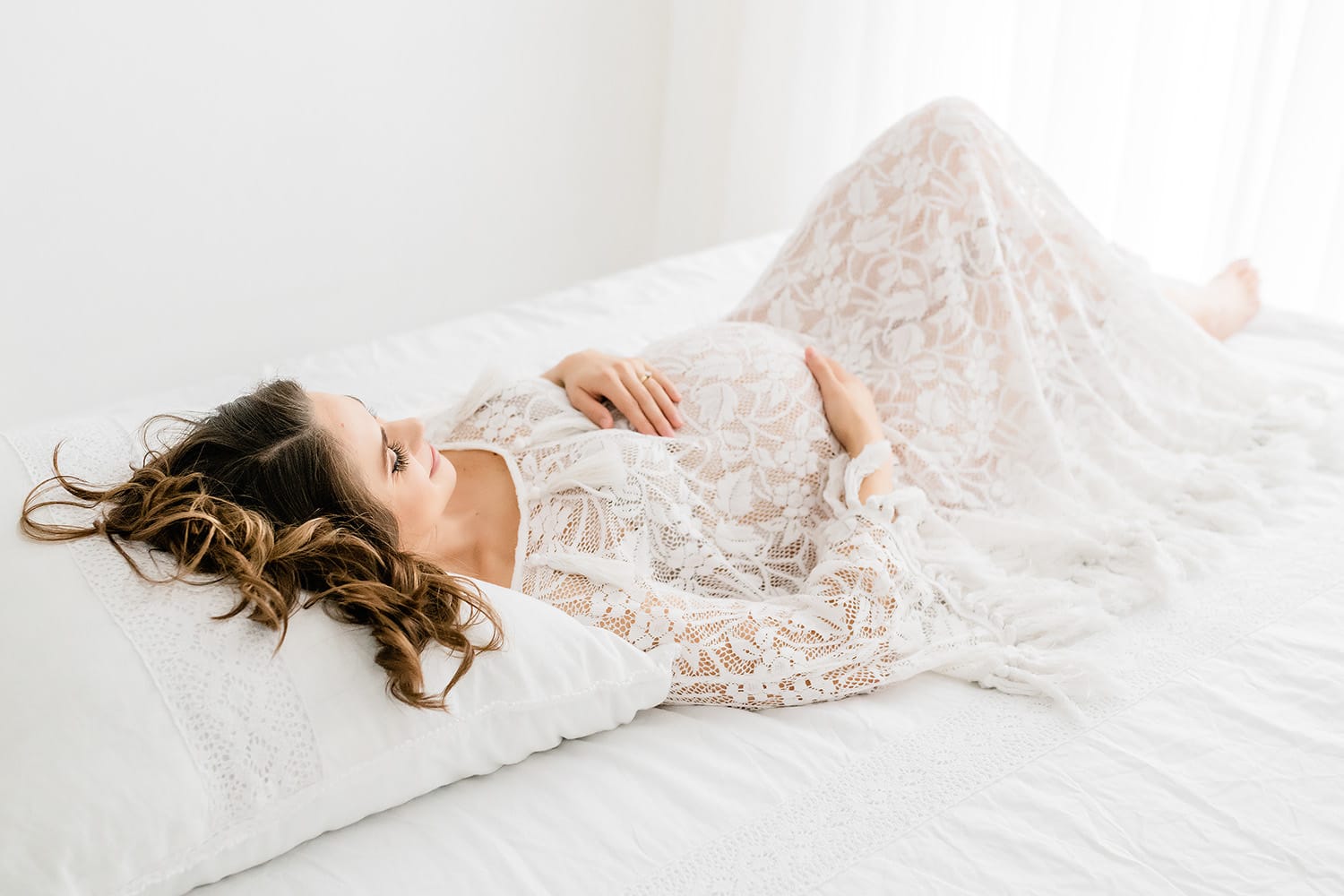 niceville-maternity-studio-portrait-white-lace-gown-reclining.jpg Alt text: Expecting mother in a sheer white lace fringe gown reclining on white bedding during an intimate studio maternity portrait in Niceville Florida