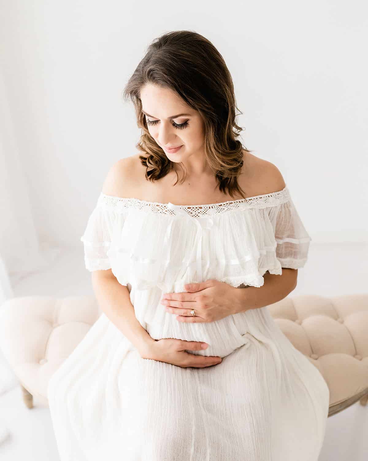 Pregnant woman in a white off-shoulder lace dress seated on a cream tufted settee cradling her bump during a studio maternity portrait in Niceville FL