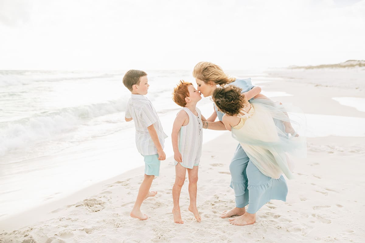 Mother leaning in to kiss her toddler son while siblings look on during a beach session on the Emerald Coast