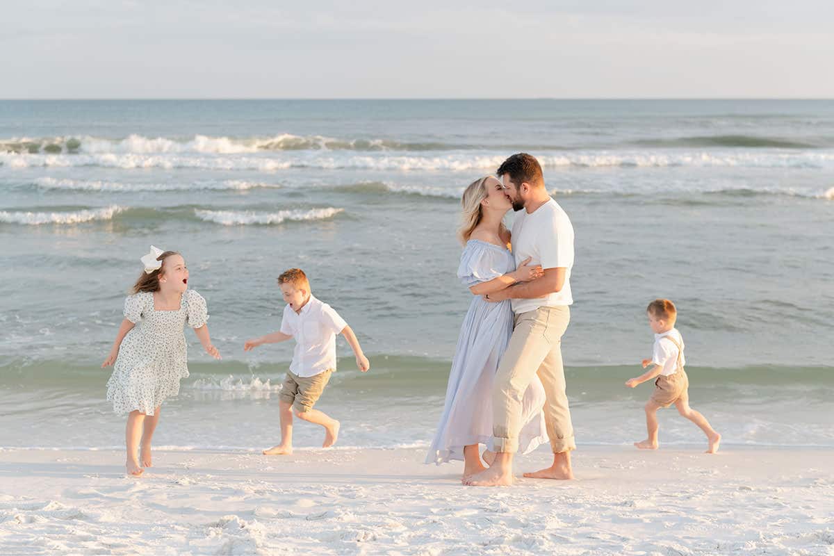 Parents sharing a kiss on the beach while their three children run and play during a Destin family photography session