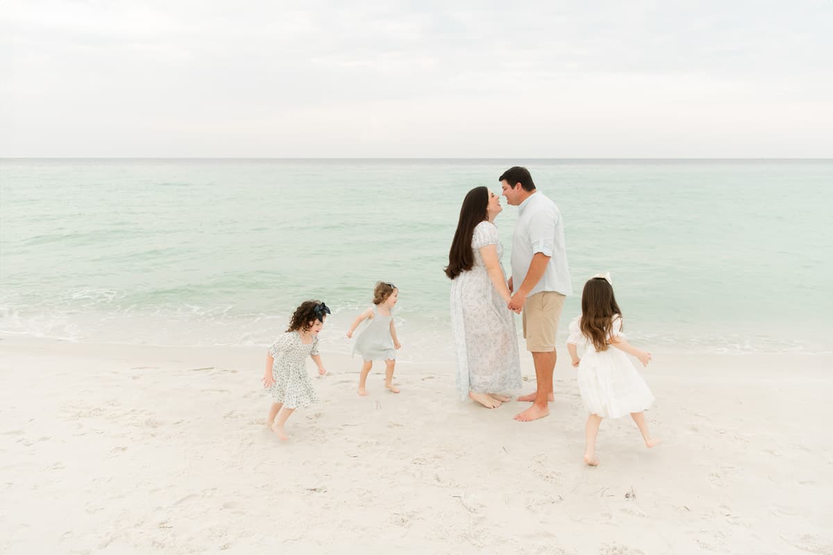 A family with three daughters walks along a Destin beach, each child in a different but complementary sage, white, or floral dress.