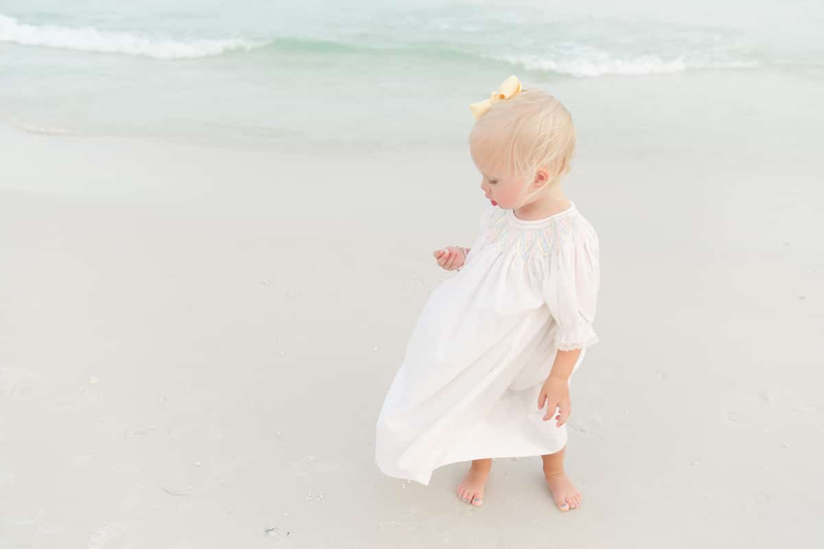 A toddler in a classic white smocked heirloom dress walks along a Destin beach during her photo session.