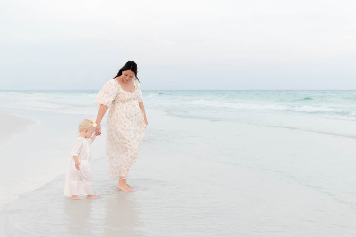 A mother and toddler walk along the shoreline in Destin wearing matching cream floral dresses for their maternity session.