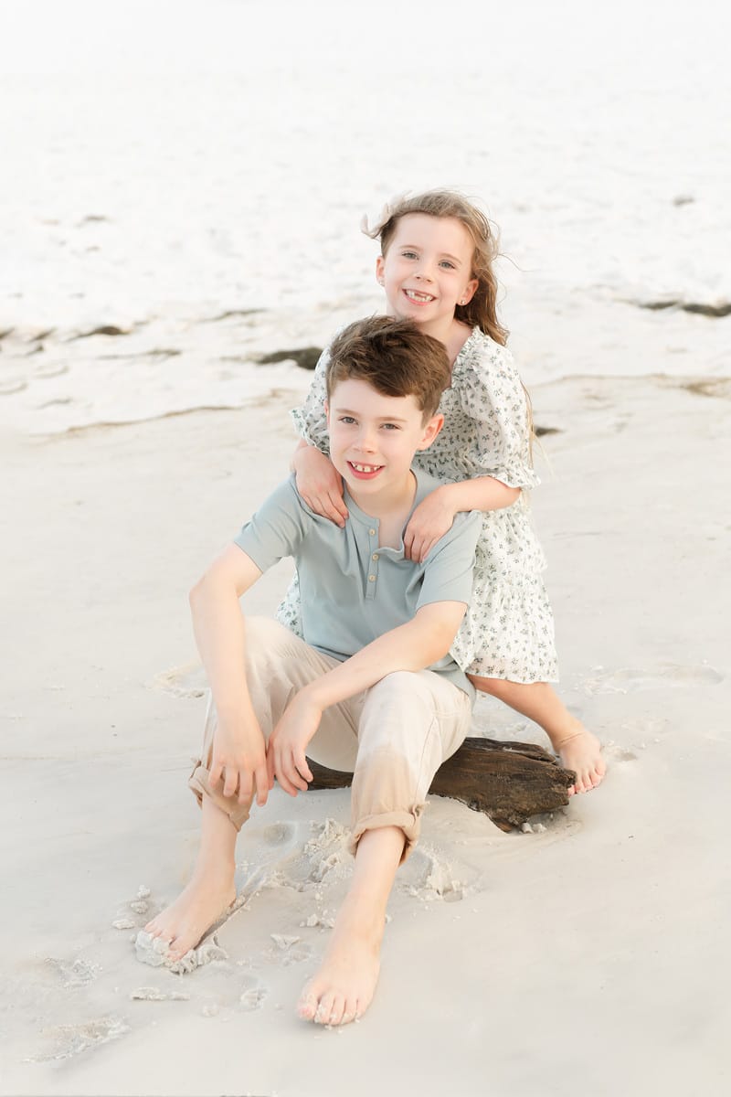 A young boy in kids clothing from stores in Niceville, FL sits on a driftwood log at the beach, smiling with a girl behind him.