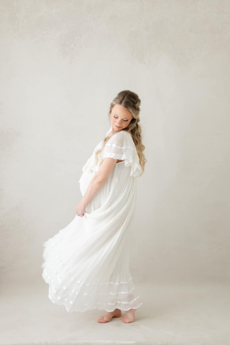 Pregnant woman in a flowing white dress, barefoot, stands softly lit&mdash;ideal shot for a book Destin maternity photographer.