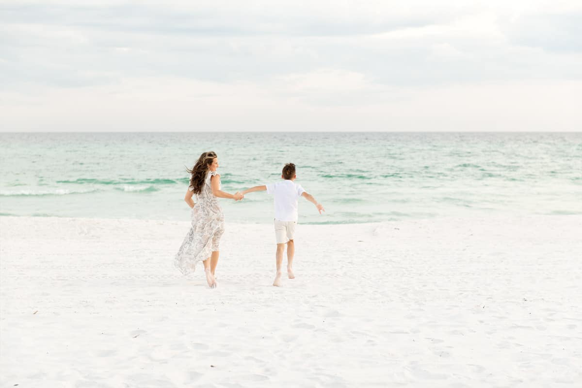 Mom in a floral dress running barefoot on the beach with her son toward the emerald Gulf water, candid and carefree moment during a Destin family session