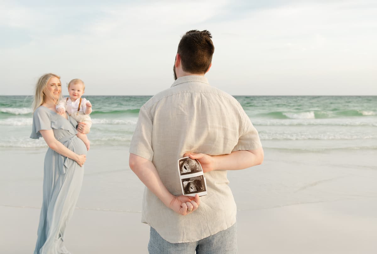Dad facing away from the camera holding ultrasound photos behind his back while mom holds their baby and smiles at him on a Destin beach, soft golden light