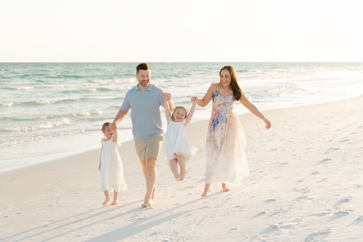 A family of four enjoys a walk on a sandy beach; parents swing one child while the other walks beside them, all smiling.