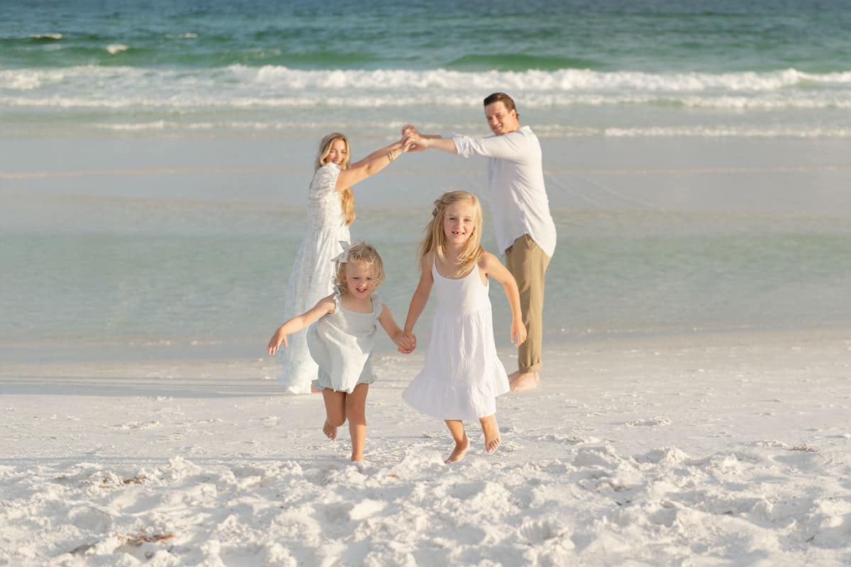 Two young sisters in white dresses running hand-in-hand on the beach while their parents form an arch overhead, golden hour light on white sand in Destin