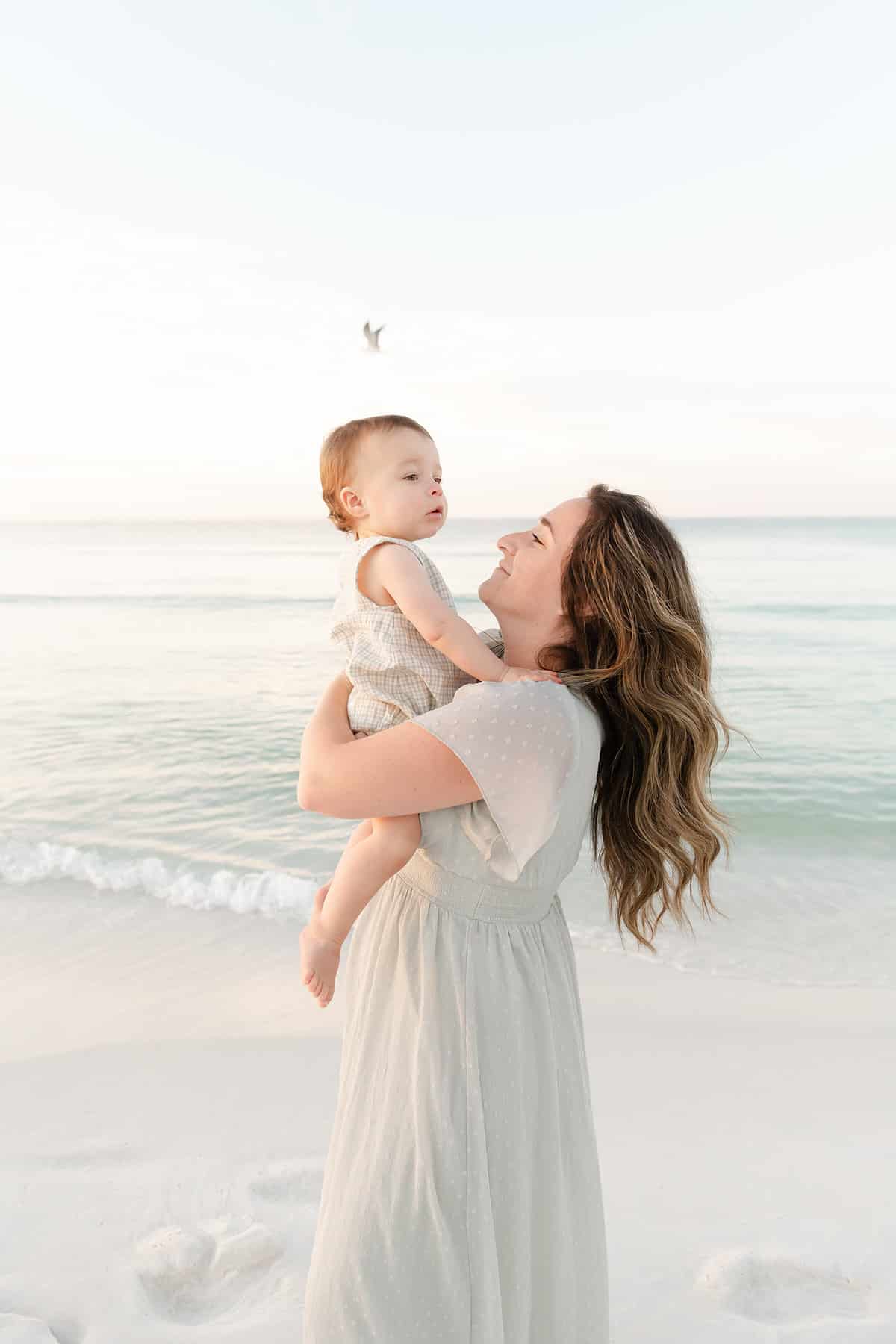 Woman holding a baby on a sandy beach near the ocean, with a bird flying in the background under a clear sky.