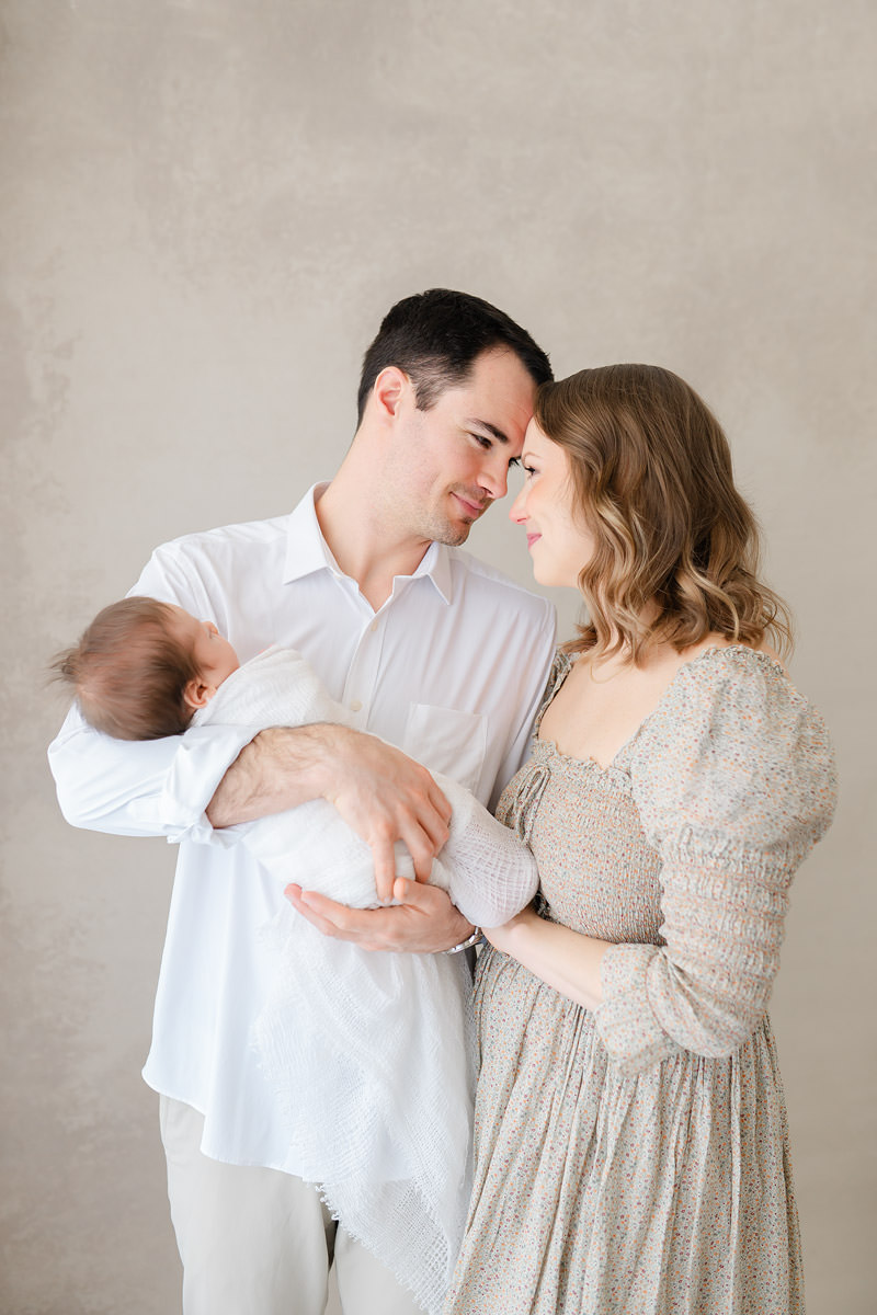 Emerald Coast newborn photographer captures mom and dad touching foreheads while dad holds their swaddled newborn in a Niceville studio